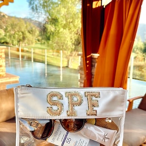 May include: A clear plastic zippered pouch with a white background and gold glitter letters spelling "SPF". The pouch contains various sunscreen bottles and tubes, including Neutrogena Ultra Sheer Dry-Touch Sunscreen SPF 55.
