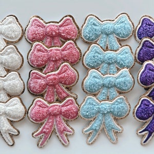 May include: A set of 15 sparkly fabric bows in white, pink, blue, and purple. The bows are all the same size and shape, and they have a gold glitter trim.
