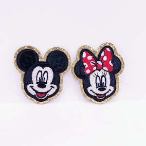 May include: Two embroidered iron-on patches featuring Mickey Mouse and Minnie Mouse. Mickey Mouse is wearing his signature red shorts and yellow shoes. Minnie Mouse is wearing a red polka dot bow and a red dress. Both patches have a gold glitter outline.