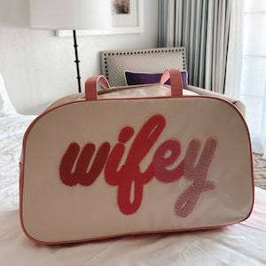 May include: A pink and white travel bag with the word "wifey" embroidered in pink and red.