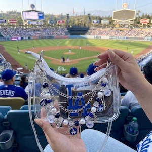 May include: Clear, transparent handbag with a blue and white Los Angeles Dodgers logo. The bag is decorated with silver chains, blue and white beads, and Dodgers charms. The background shows a baseball stadium.