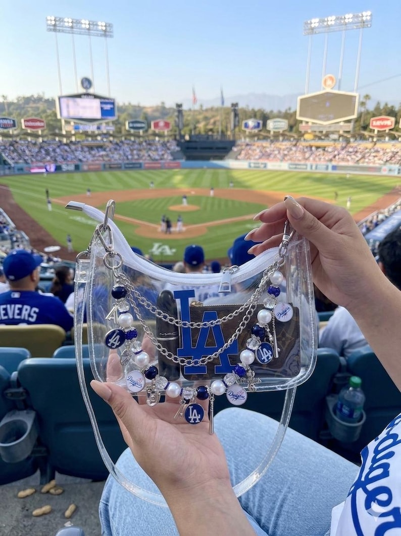 May include: Clear, transparent handbag with a blue and white Los Angeles Dodgers logo. The bag is decorated with silver chains, blue and white beads, and Dodgers charms. The background shows a baseball stadium.
