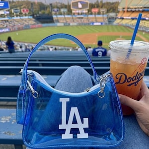 May include: A clear blue handbag with the LA Dodgers logo in white. The bag has a matching blue handle and silver hardware. A clear plastic cup with a blue straw and the word "Dodgers" is also visible.