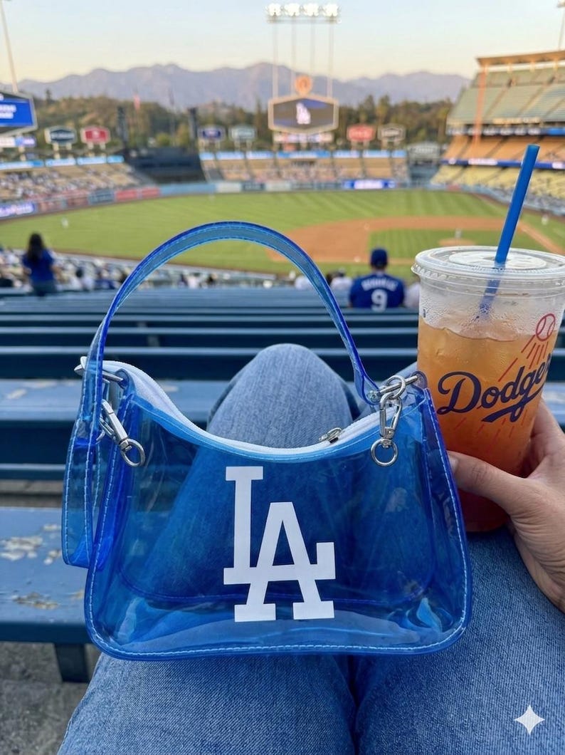 May include: A clear blue handbag with the LA Dodgers logo in white. The bag has a matching blue handle and silver hardware. A clear plastic cup with a blue straw and the word "Dodgers" is also visible.
