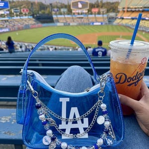 May include: A clear blue handbag with a white "LA" logo, silver chain, and beaded accents. The bag has a handle and a zipper closure. A drink with a blue straw and the Dodgers logo is visible.