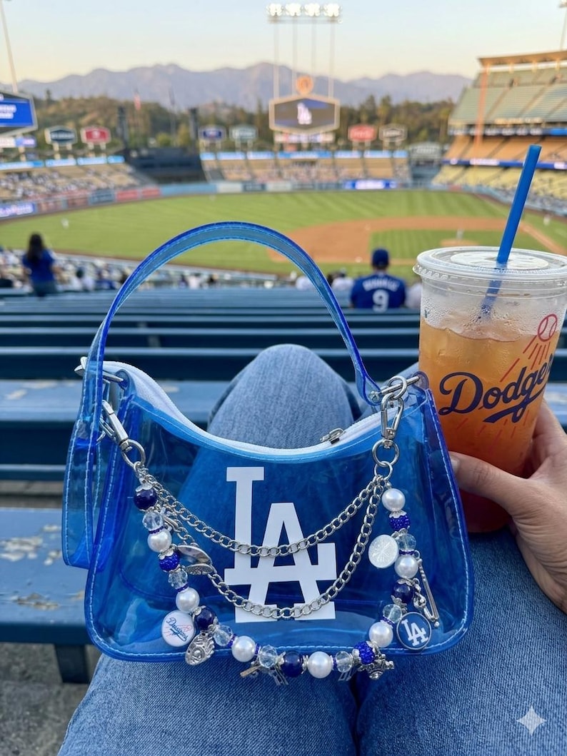 May include: A clear blue handbag with a white "LA" logo, silver chain, and beaded accents. The bag has a handle and a zipper closure. A drink with a blue straw and the Dodgers logo is visible.