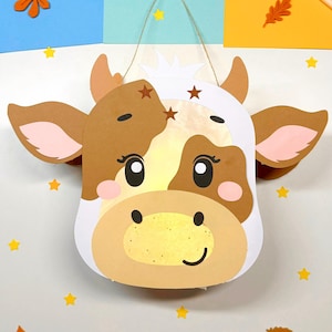 Può includere: Una lanterna di carta a forma di mucca marrone e bianca con un naso giallo e orecchie rosa. La lanterna è appesa a una corda ed è circondata da stelle gialle e foglie arancioni.