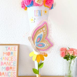 Pode incluir: Um cone escolar branco decorado com flores de papel coloridas, uma borboleta e o nome "LENI". A bolsa é adornada com um laço amarelo e uma base de flor de papel. Uma impressão emoldurada com o texto "TAKE MORE CHANCES DANCE MORE DANCES" é visível.