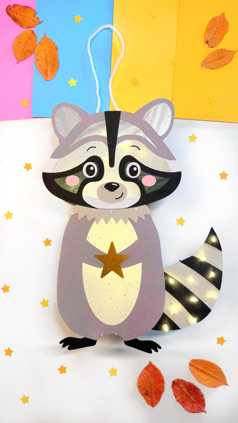 May include: A gray raccoon paper lantern with a black nose and eyes, a gold star, and a striped tail. The lantern has a white string for hanging and is decorated with small yellow stars.