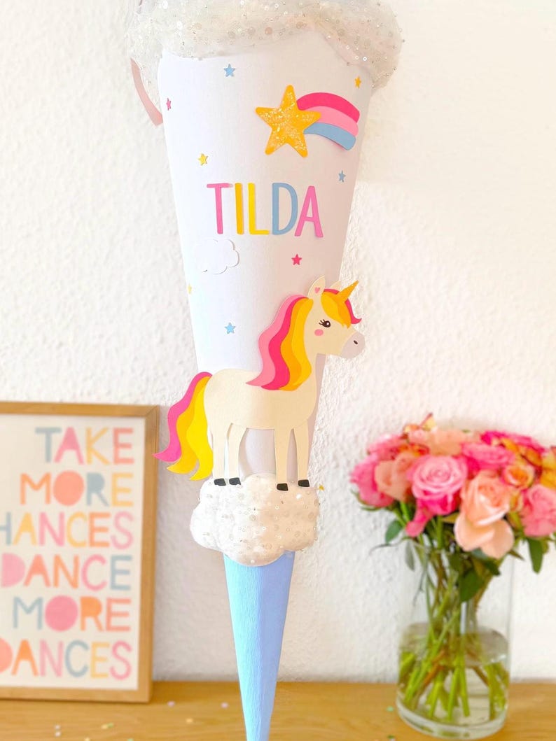May include: A white paper cone with a unicorn, a rainbow, and a star. The cone is decorated with glitter and has the name "Tilda" written on it.