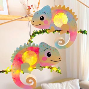 Könnte beinhalten: Zwei bunte Chamäleon-Laternen mit Regenbogen-Farbverlauf, die auf einem Ast sitzen. Jede Laterne hat ein gelbes Leuchtmittel und ist mit grünen Blättern und kleinen Lichtern verziert. Das Design ist verspielt und farbenfroh.