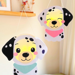 Könnte beinhalten: Zwei hängende Hundelaternen. Jede Laterne zeigt ein lächelndes Hundegesicht mit schwarzen Flecken, schwarzen Ohren und einem bunten Halstuch. Die Laternen sind von innen beleuchtet und erzeugen ein warmes Leuchten.