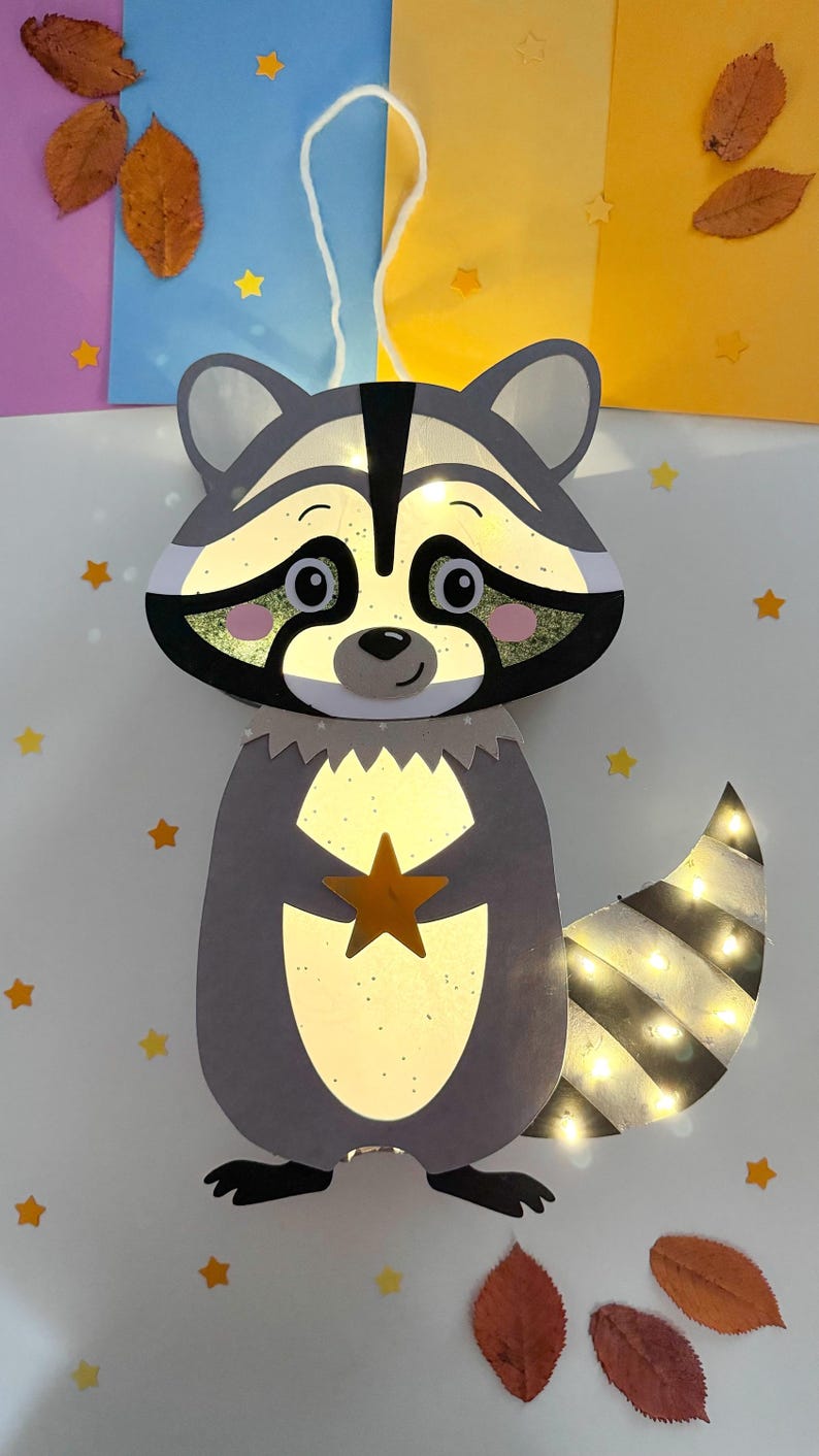 May include: A gray raccoon paper lantern with a white string for hanging. The raccoon is holding a gold star and has a striped tail with lights. The lantern is surrounded by yellow stars and brown leaves on a white background.
