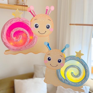 Peut inclure: Deux lampes décoratives en forme d'escargot, idéales pour une chambre d'enfant. L'une a une coquille rose, l'autre bleue, toutes deux avec un motif en spirale. Les escargots ont des visages souriants et des étoiles.