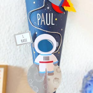 Puede incluir: Un cono escolar azul y plateado decorado con un cohete, un astronauta y el nombre "PAUL". El cono tiene un dise&ntilde;o de cielo estrellado y un letrero que dice "1. Klasse".