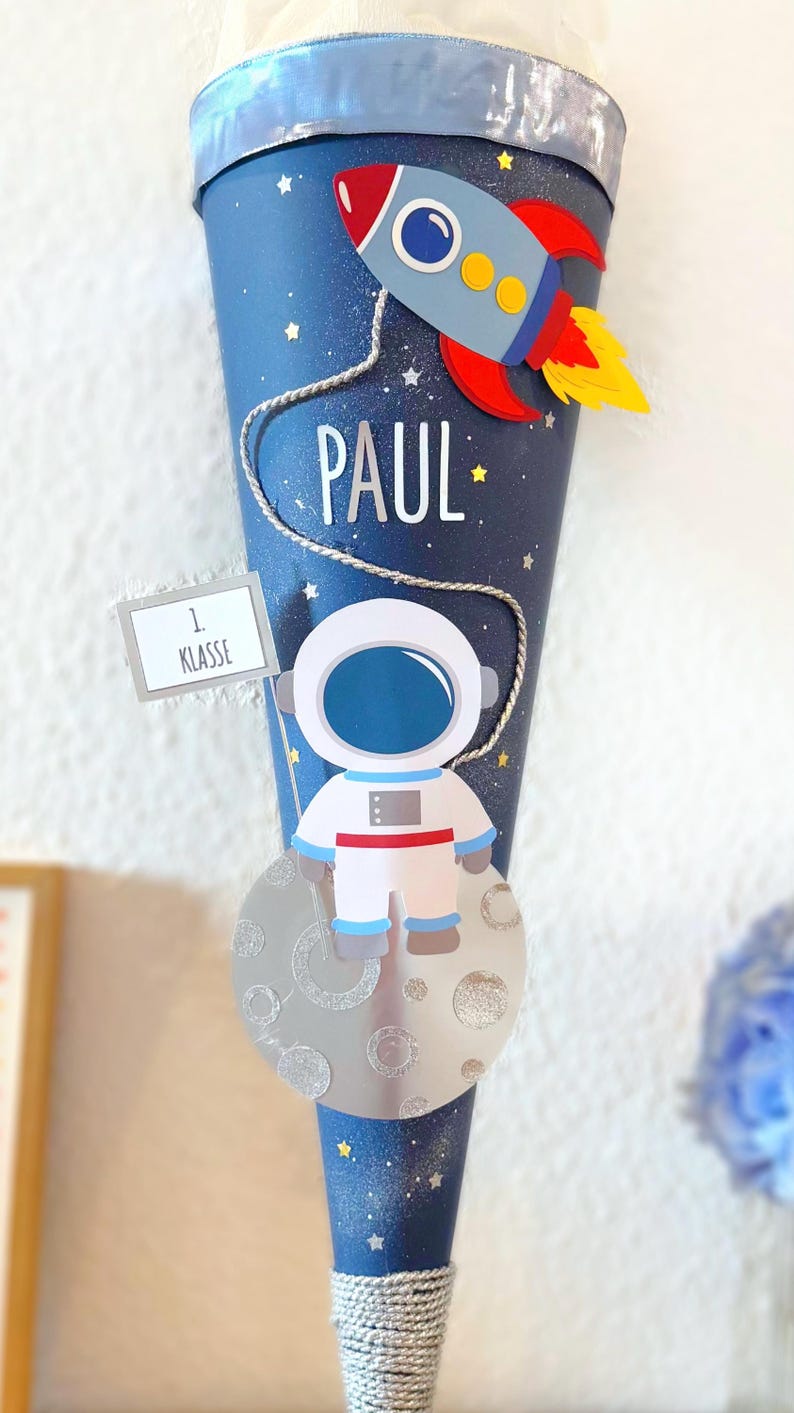 Puede incluir: Un cono escolar azul y plateado decorado con un cohete, un astronauta y el nombre "PAUL". El cono tiene un dise&ntilde;o de cielo estrellado y un letrero que dice "1. Klasse".