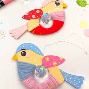 K&ouml;nnte beinhalten: Zwei handgefertigte Vogelornamente. Jeder Vogel hat einen gelben K&ouml;rper, einen mit rotem oder rosa Garn umwickelten Bauch und blaue oder rosa Fl&uuml;gel mit Punkten. In der Mitte jedes Vogels befindet sich eine Kristallperle.