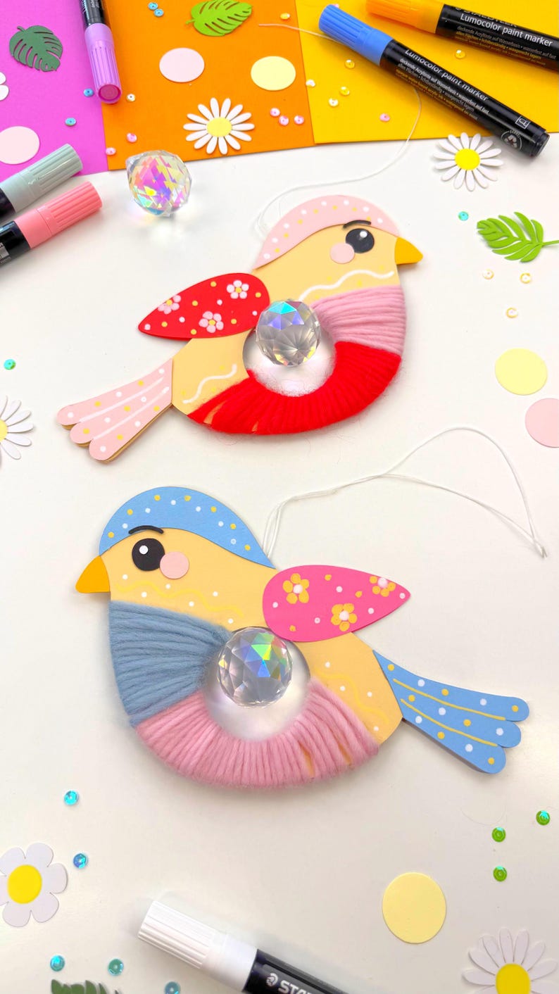 K&ouml;nnte beinhalten: Zwei handgefertigte Vogelornamente. Jeder Vogel hat einen gelben K&ouml;rper, einen mit rotem oder rosa Garn umwickelten Bauch und blaue oder rosa Fl&uuml;gel mit Punkten. In der Mitte jedes Vogels befindet sich eine Kristallperle.