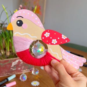 K&ouml;nnte beinhalten: Ein verspieltes, vogelartiges Bastelobjekt mit einem Kristallanh&auml;nger. Der Vogel hat einen rosa-beigen K&ouml;rper, rote Fl&uuml;gelelemente mit wei&szlig;en Blumen und eine rote Garnbasis. F&uuml;r Dekoration oder kreative Projekte.
