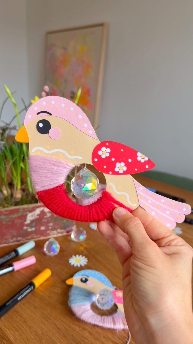 K&ouml;nnte beinhalten: Ein verspieltes, vogelartiges Bastelobjekt mit einem Kristallanh&auml;nger. Der Vogel hat einen rosa-beigen K&ouml;rper, rote Fl&uuml;gelelemente mit wei&szlig;en Blumen und eine rote Garnbasis. F&uuml;r Dekoration oder kreative Projekte.
