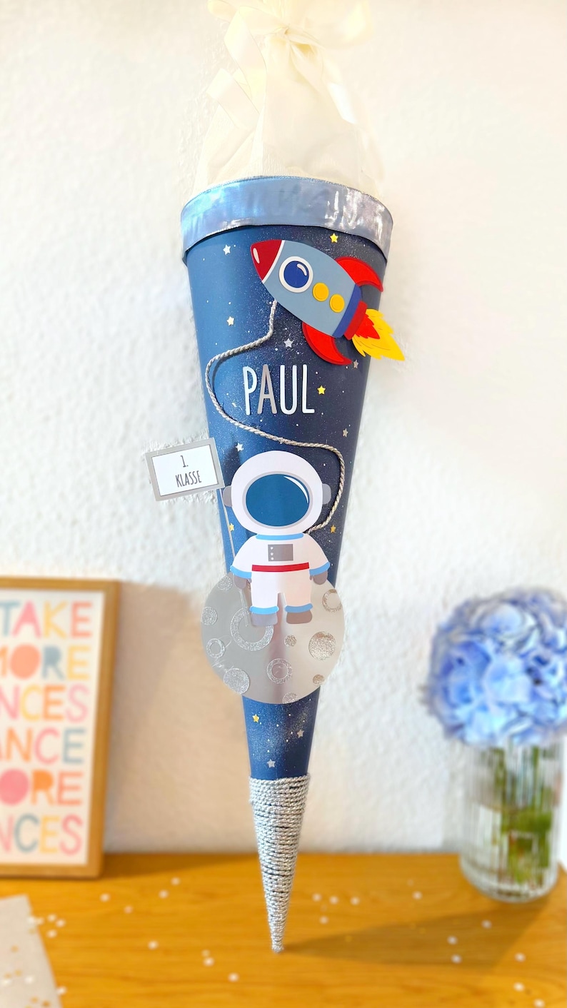 Puede incluir: Un cono escolar azul decorado con un astronauta, un cohete y el nombre "PAUL". La bolsa tiene una base de cuerda plateada y est&aacute; rematada con una cinta blanca. Un peque&ntilde;o letrero dice "1 Klasse".