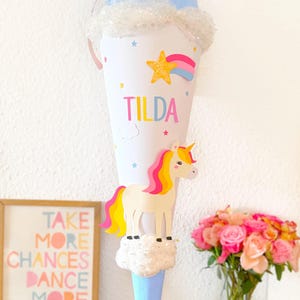 May include: A blue and white unicorn school cone with the name "Tilda" written on it. The cone is decorated with stars, a rainbow, and a cloud. The cone is sitting on a wooden table with a picture frame and a vase of pink roses.