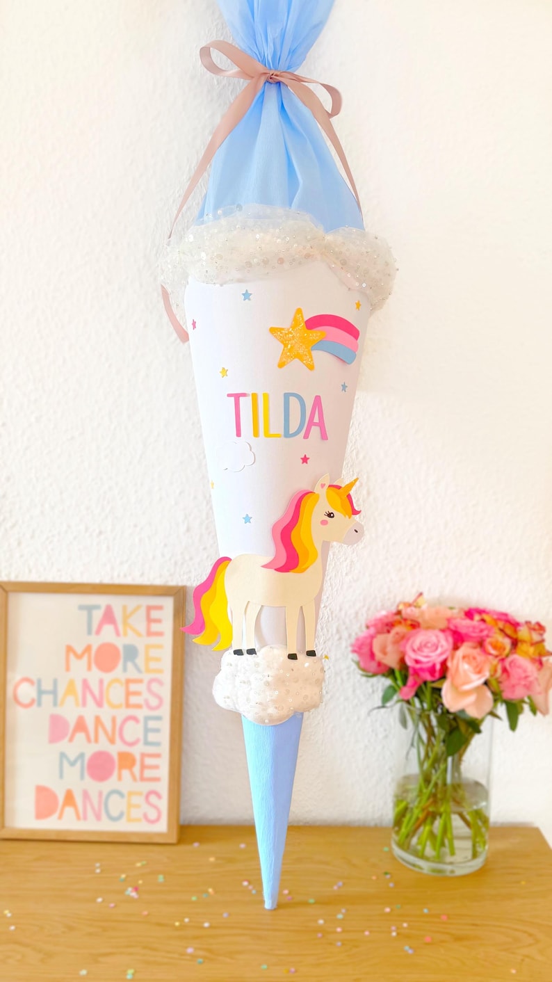 May include: A blue and white unicorn school cone with the name "Tilda" written on it. The cone is decorated with stars, a rainbow, and a cloud. The cone is sitting on a wooden table with a picture frame and a vase of pink roses.