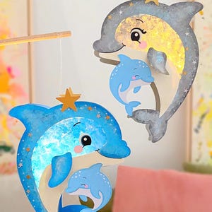 Peut inclure: Deux décorations suspendues en forme de dauphins accrochées à des tiges en bois. Un dauphin est bleu avec une finition pailletée et un petit, tandis que l'autre est gris avec un ventre doré pailleté et un petit. Tous deux ont des étoiles dorées.