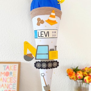 Op de afbeelding: Een witte, kegelvormige schooltas versierd met een geel en blauw bouwthema. De tas heeft een gele graafmachine, een verkeerskegel en de naam "LEVI" met een datum. De bovenkant is blauw met gele linten.