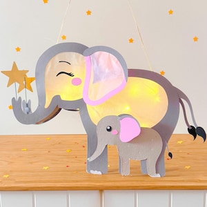 May include: A grey and pink paper elephant with a smaller grey and pink elephant inside. The larger elephant is holding a gold star. The elephants are lit up with white lights.