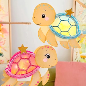 Puede incluir: Dos decoraciones de tortugas colgantes con caparazones iluminados. Una tortuga tiene un caparazón azul con centro amarillo y una estrella dorada, la otra tiene un caparazón rosa con centro de luz cálida y una estrella dorada. Piel naranja claro.