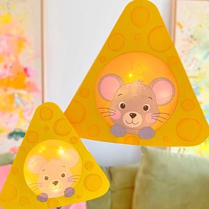 Peut inclure: Deux lanternes triangulaires jaunes en forme de quartiers de fromage, chacune dotée d'une découpe circulaire avec une illustration de visage de souris. Les lanternes sont éclairées de l'intérieur, diffusant une lueur chaude par les ouvertures.