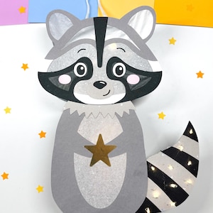May include: A gray raccoon paper lantern with a black and white striped tail. The lantern has a gold star on its chest and is decorated with white glitter. The lantern is hanging from a white string and is surrounded by yellow stars and orange leaves.
