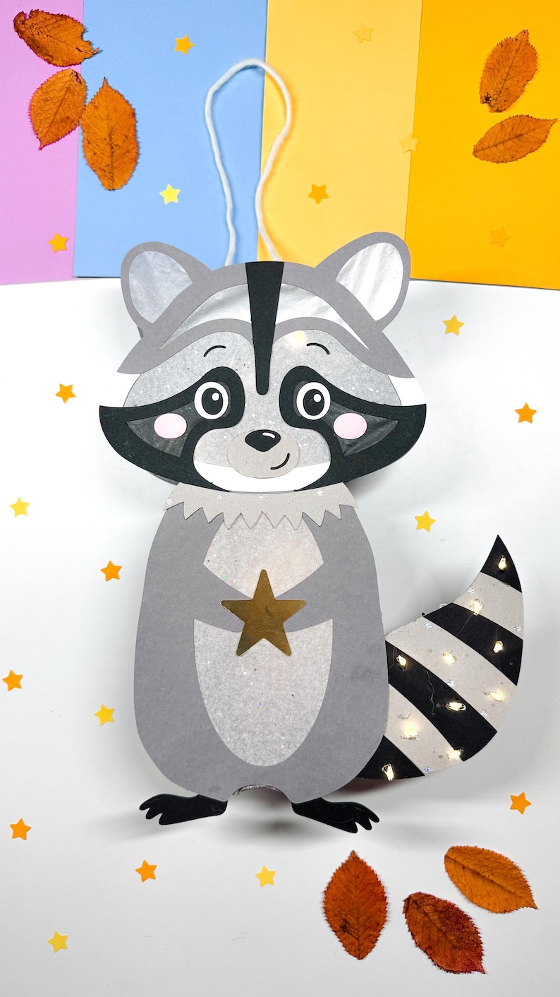 May include: A gray raccoon paper lantern with a black and white striped tail. The lantern has a gold star on its chest and is decorated with white glitter. The lantern is hanging from a white string and is surrounded by yellow stars and orange leaves.