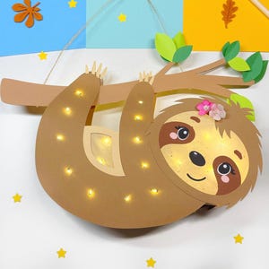 May include: A brown sloth with a pink flower on its head hangs from a brown branch with green leaves. The sloth is made of paper and has small white lights inside. The background is a white surface with yellow stars and brown leaves.