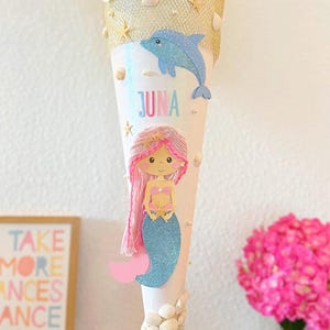 Può includere: Un cono scolastico bianco a forma di cono decorato con un tema sirena e delfino. La borsa presenta una sirena con capelli rosa e coda blu, un delfino blu e il nome "JUNA". La parte superiore è adornata con tessuto dorato e conchiglie.