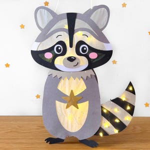 Puede incluir: Una linterna de papel con forma de mapache gris con una cola rayada en blanco y negro. La linterna tiene una estrella dorada en el pecho y está iluminada con pequeñas luces blancas.