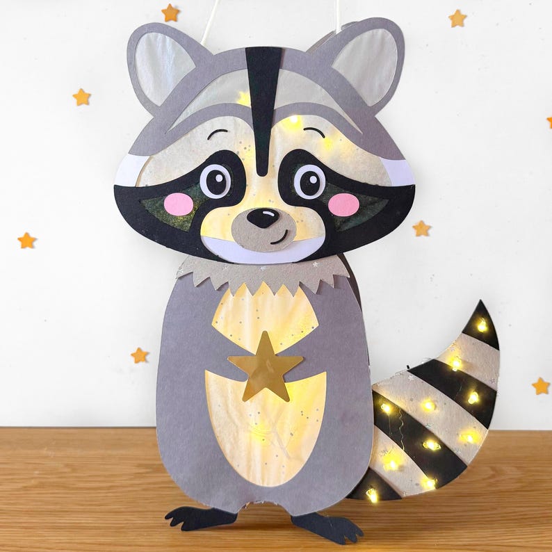 May include: A gray raccoon-shaped paper lantern with a black and white striped tail. The lantern has a gold star on its chest and is lit with small white lights.