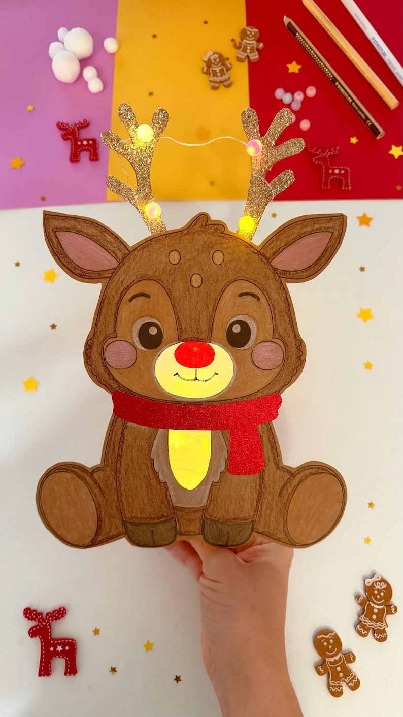 May include: A brown, cartoon-style reindeer decoration with a red scarf and a glowing yellow belly and nose. The reindeer has glittery antlers with small lights. The image includes gingerbread men and star-shaped confetti, suggesting a Christmas theme.