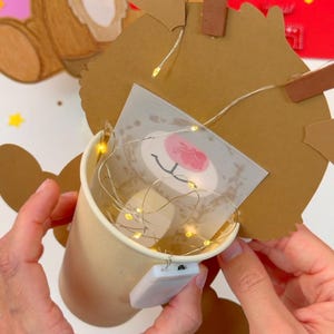 May include: A handmade Christmas decoration featuring a reindeer design. The reindeer is crafted from brown cardboard with a printed face and a red nose. A small paper cup holds fairy lights, creating a warm glow. The decoration is held by hands.
