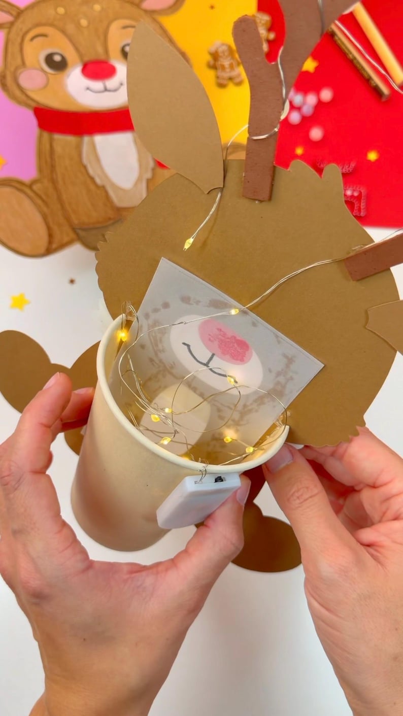 May include: A handmade Christmas decoration featuring a reindeer design. The reindeer is crafted from brown cardboard with a printed face and a red nose. A small paper cup holds fairy lights, creating a warm glow. The decoration is held by hands.