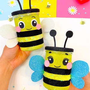 Puede incluir: Dos tarros artesanales con temática de abeja, con relleno de purpurina amarilla, rayas negras y caras de dibujos animados. Tienen antenas negras, mejillas rosas y alas brillantes. Los tarros se sostienen sobre un fondo colorido con materiales de manualidades.