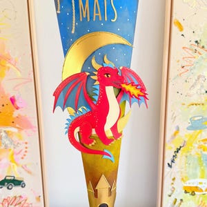 Può includere: Un contenitore regalo a forma di cono con uno sfondo di cielo notturno blu e il nome MATS in lettere dorate. Un drago rosso con ali blu sputa fuoco davanti a una luna crescente dorata. La base presenta una sagoma di castello dorato e finiture.