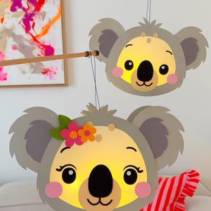 Pode incluir: Duas lanternas de urso-koala com rostos amarelos brilhantes, orelhas cinzentas e bochechas rosadas estão suspensas em varas de madeira. Uma lanterna tem uma coroa de flores. As lanternas estão penduradas em frente a uma parede branca e uma pintura abstrata colorida.