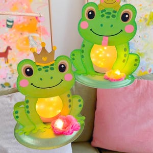 May include: Two hanging frog decorations featuring green bodies, pink cheeks, and illuminated bellies. One frog wears a gold crown, while the other has a small frog on its head. Each sits on a green base with a pink flower and warm internal light.