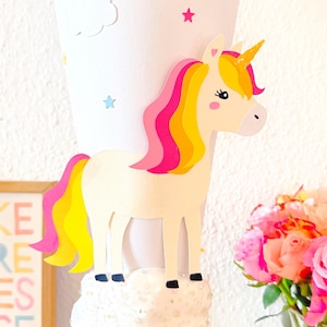 May include: A white paper cone decorated with a unicorn, stars, and clouds. The cone has the name "Tilda" written on it in pink, yellow, and blue letters.