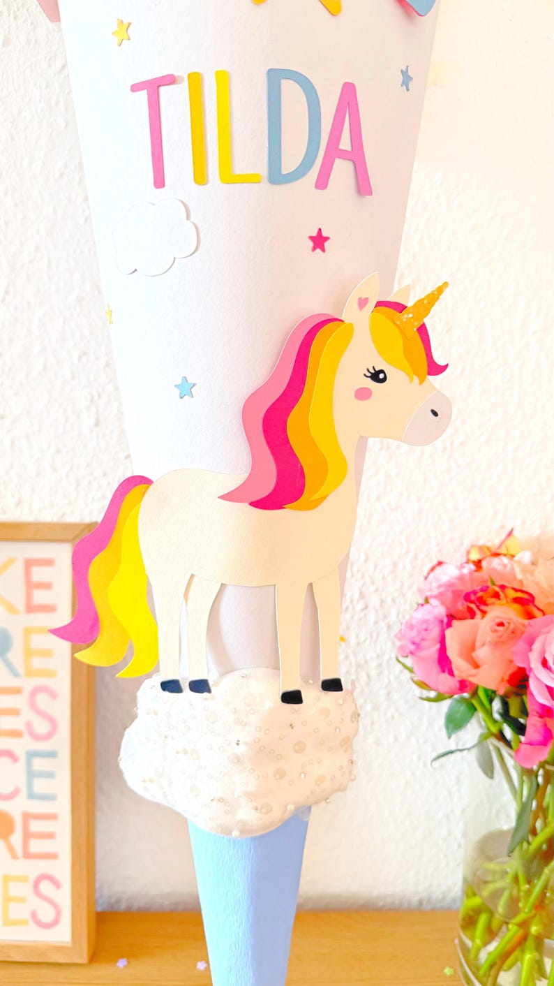 May include: A white paper cone decorated with a unicorn, stars, and clouds. The cone has the name "Tilda" written on it in pink, yellow, and blue letters.