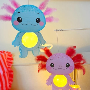 Può includere: Due lanterne di axolotl stravaganti. Uno è azzurro con accenti di piume rosa, l'altro è lavanda con accenti di piume rosse. Entrambi hanno una luce gialla al centro e sono sospesi a un'asta di legno.