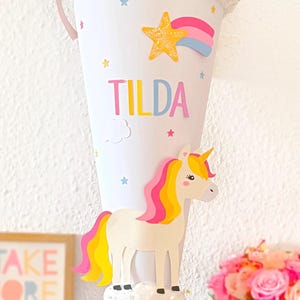 May include: A blue and white cone-shaped school bag with a unicorn, rainbow, and stars. The bag is decorated with glitter and has the name "TILDA" written on it.