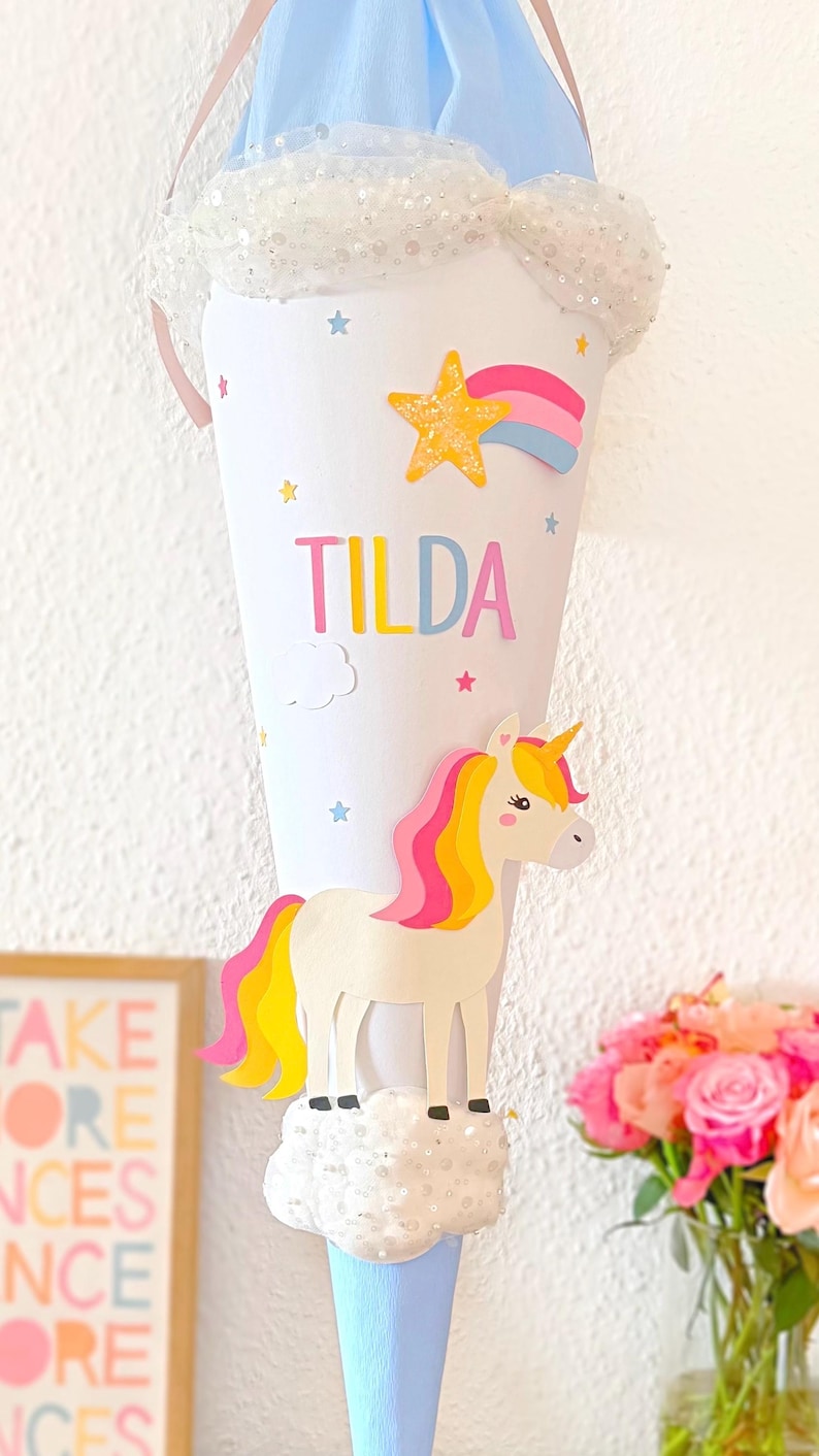 May include: A blue and white cone-shaped school bag with a unicorn, rainbow, and stars. The bag is decorated with glitter and has the name "TILDA" written on it.